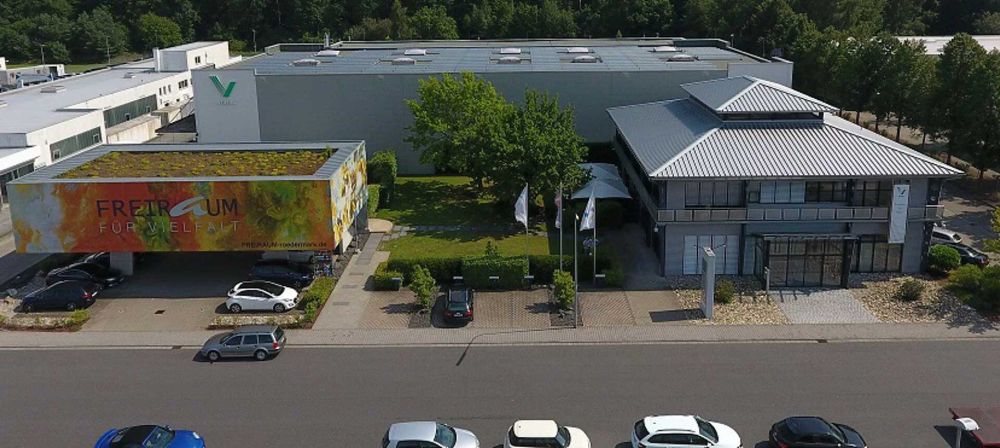Luftaufnahme eines modernen Firmengebäudes mit Parkplatz und angrenzenden Hallen als Symbol für Unternehmensstandort, Infrastruktur und Betriebsflächen