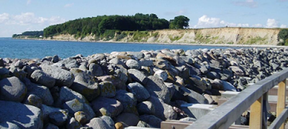 Küstenlandschaft mit Steinufer, Meer und bewaldeter Steilküste unter blauem Himmel als ruhige Naturszene